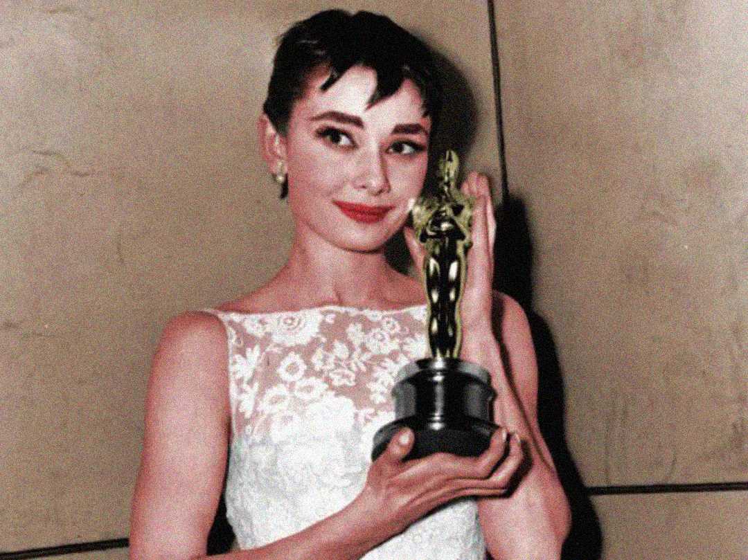 A woman in a white lace dress holds an Oscar statuette and smiles, standing in front of a plain tan background.