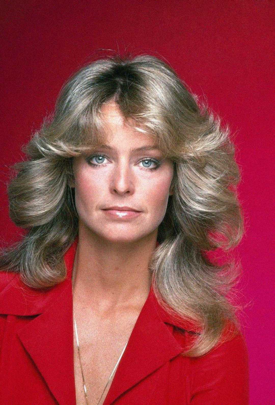 A woman with voluminous, feathered blonde hair and blue eyes is wearing a red collared top, posing against a solid pink-red background. She has a neutral expression and minimal jewelry.