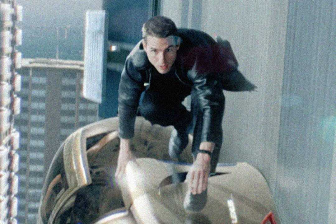 A man in a black jacket is crouched on top of a speeding car driving vertically down the side of a glass skyscraper, with city buildings blurred in the background.