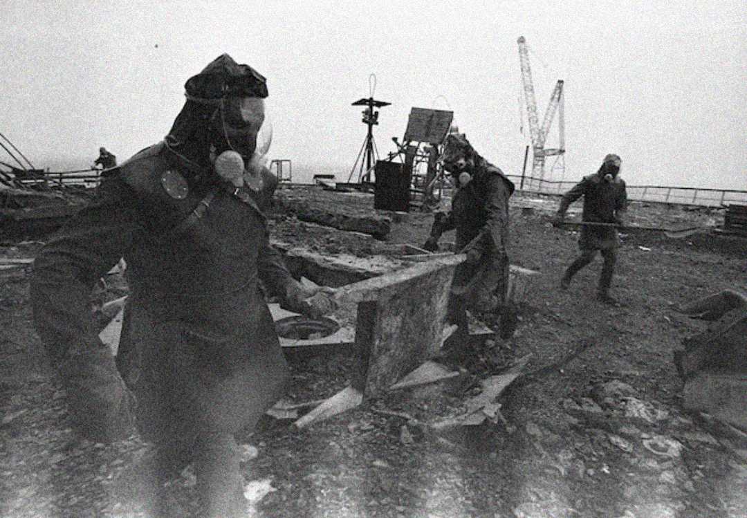 Three people wearing gas masks and protective suits work amid debris on a devastated, industrial site. The background is foggy, with cranes and damaged equipment visible, creating a tense, hazardous atmosphere.