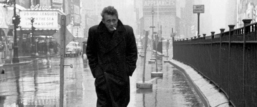 A man in a long coat walks alone on a wet city street, hands in his pockets, with buildings and fog in the background. He appears thoughtful as he smokes a cigarette.