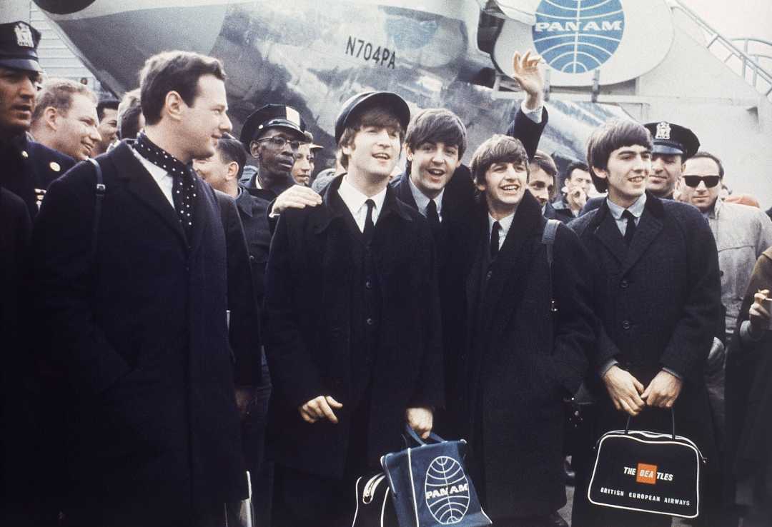 Four young men in suits stand smiling and waving in front of a Pan Am airplane, surrounded by a crowd. Two hold Pan Am bags, and one holds a black bag labeled “The Beatles.”