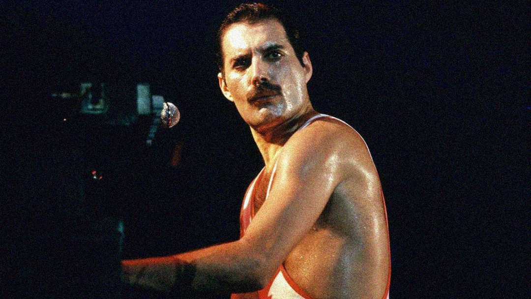 A man with short hair and a mustache is playing the piano and singing into a microphone on stage under spotlight, wearing a sleeveless top, with sweat on his face and arms.