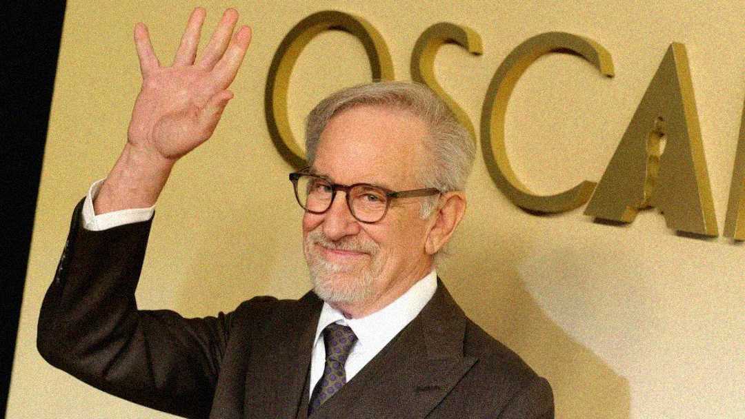 An older man with gray hair, glasses, and a beard smiles and waves while standing in front of a wall with large gold letters spelling "OSCARS." He is wearing a suit and tie.
