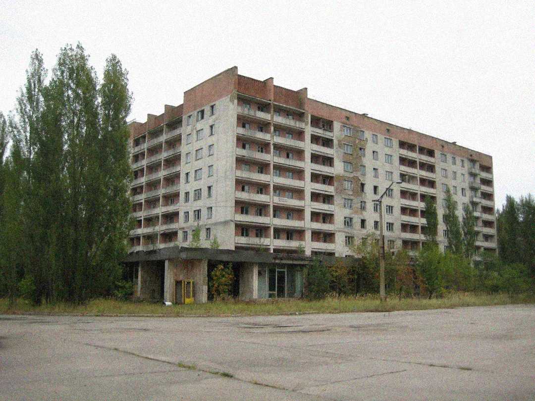 A large, abandoned apartment building with multiple floors and balconies, surrounded by overgrown vegetation and trees, stands next to an empty, cracked concrete area under an overcast sky.