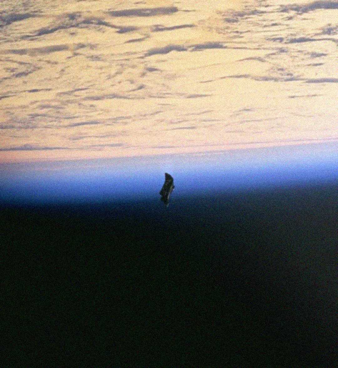 A dark object floats above Earth’s horizon, with a layer of clouds and a blue atmospheric glow visible in the background, against the blackness of space.