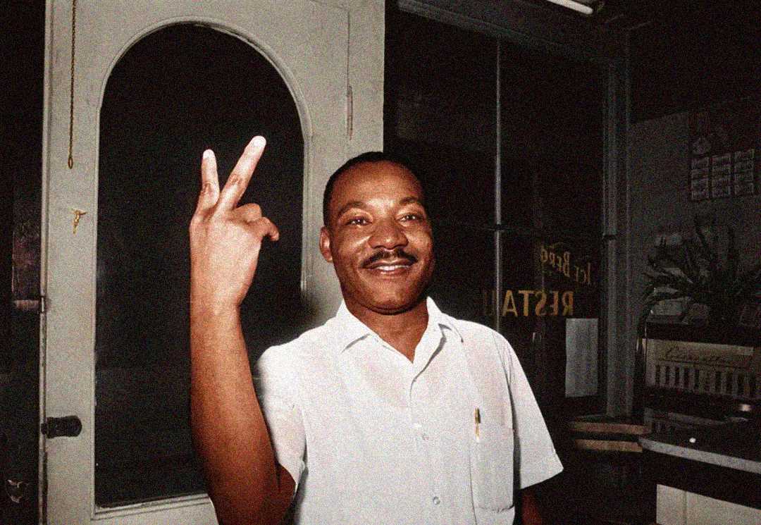 A smiling man in a short-sleeve shirt stands indoors, holding up two fingers in a peace sign. A glass-paned door and various objects are in the background.