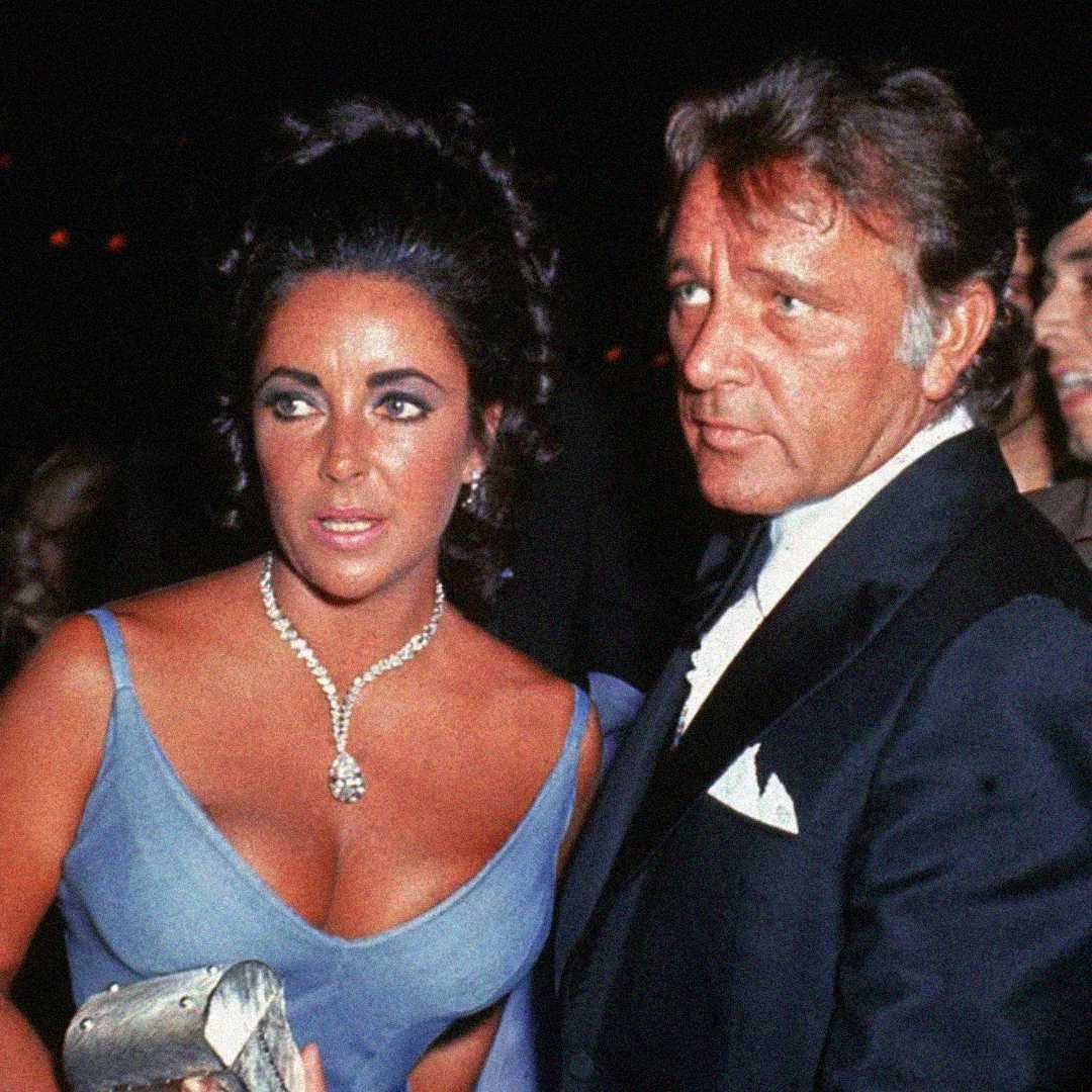 A woman in a blue dress with a diamond necklace stands next to a man in a tuxedo at a formal event. Both have dark hair and serious expressions, and the woman is holding a silver clutch.