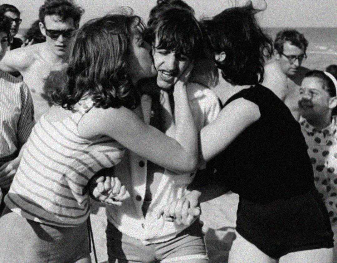 Black and white photo of a group at the beach; two women excitedly hug and kiss a smiling man in the center, surrounded by other people enjoying the sunny day.