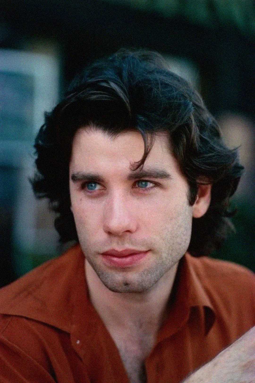 A young man with medium-length dark hair and blue eyes looks slightly to the side. He is wearing a rust-colored shirt and has a thoughtful expression on his face. The background is blurred.