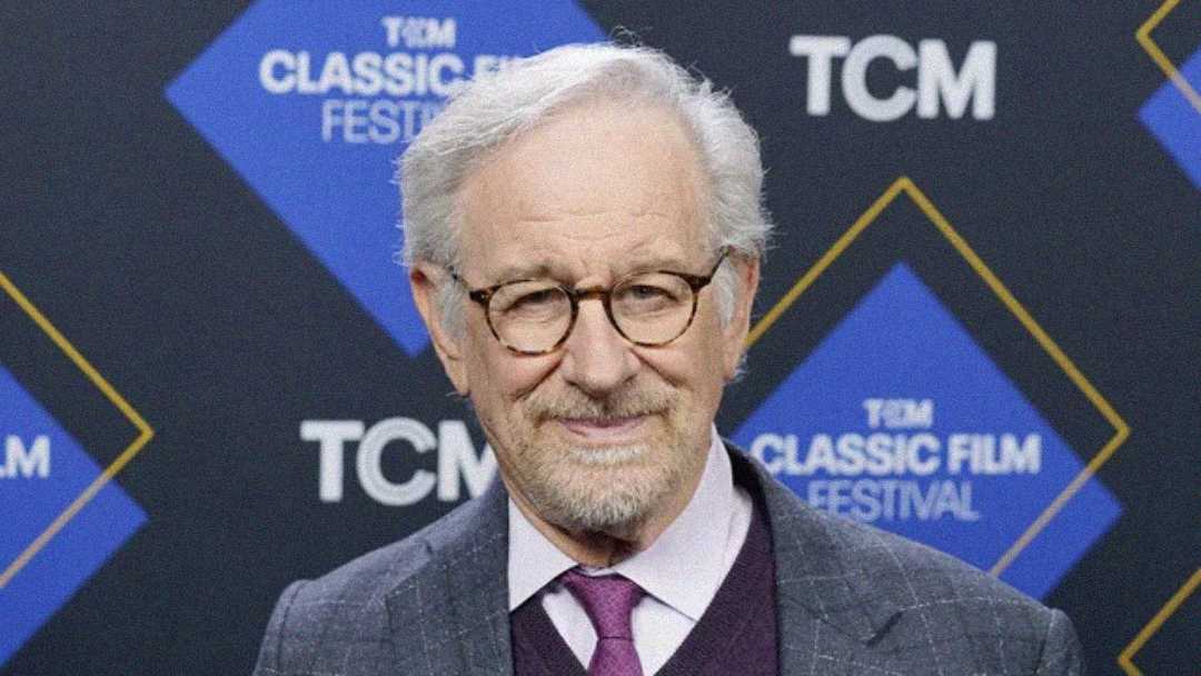 An older man with gray hair, glasses, and a beard smiles while standing in front of a blue and black backdrop with "TCM Classic Film Festival" logos.