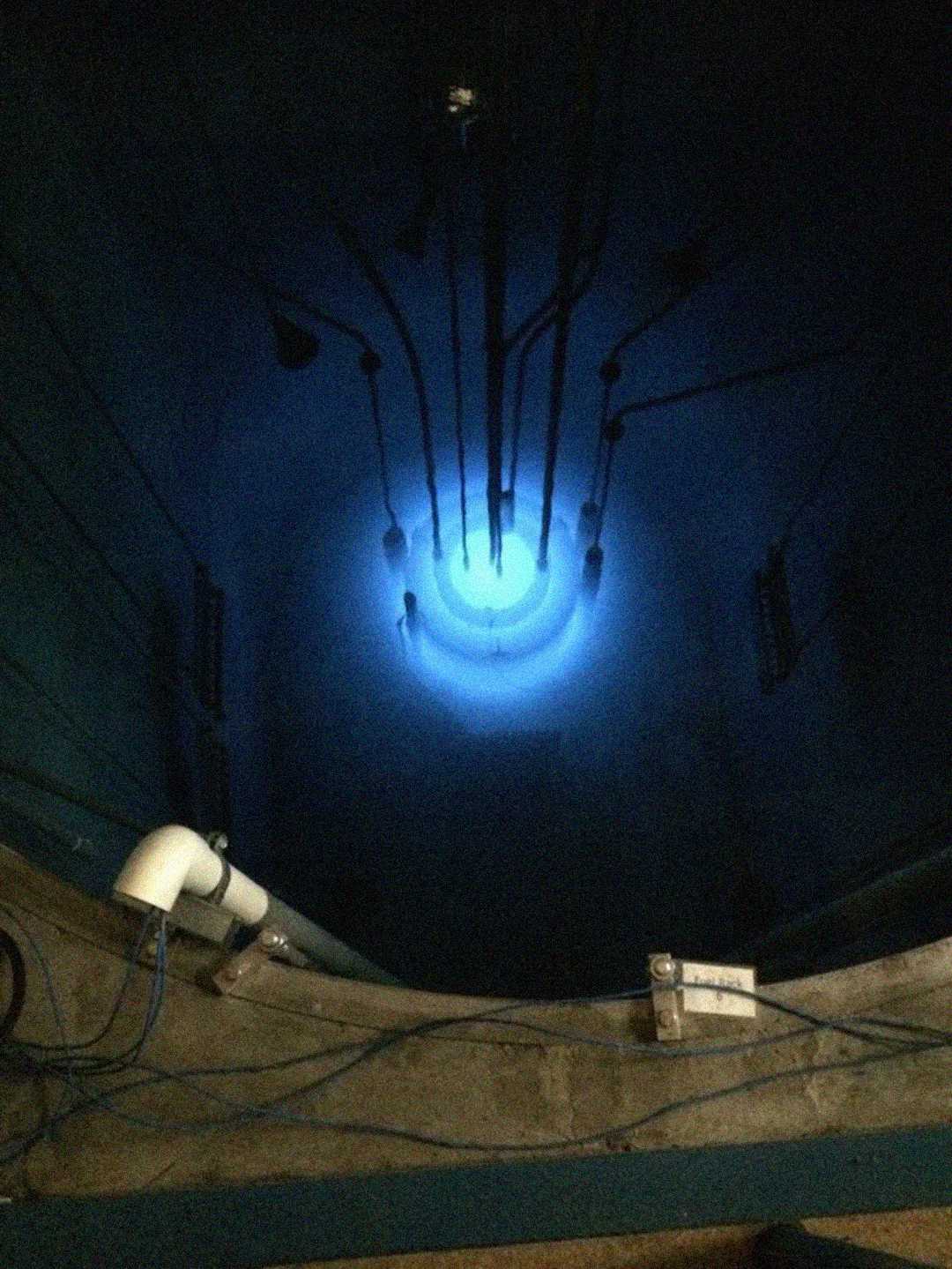 A glowing blue light, known as Cherenkov radiation, emanates from the core of a nuclear reactor submerged in water, with several rods or instruments immersed in the pool.