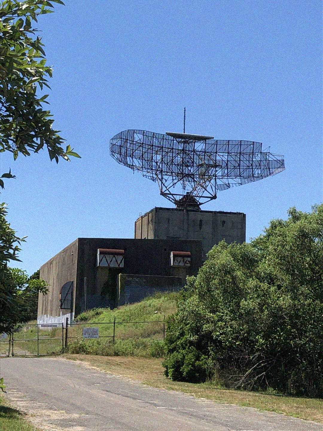 An old military radar installation sits atop a concrete building, surrounded by green trees and grass, under a clear blue sky. A paved path leads up to the fenced-off structure.