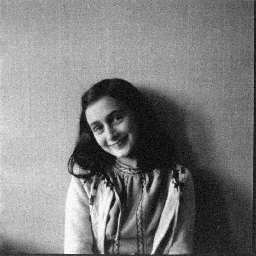 A young girl with dark hair smiles softly while sitting against a plain wall, wearing a light-colored blouse with embroidered details. The black-and-white photo has a simple, warm, and timeless feel.