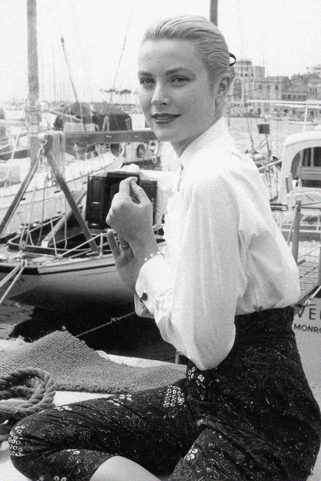 A woman with blonde hair pulled back, wearing a white blouse and patterned skirt, sits by a marina with sailboats in the background, smiling slightly at the camera in a black and white photo.