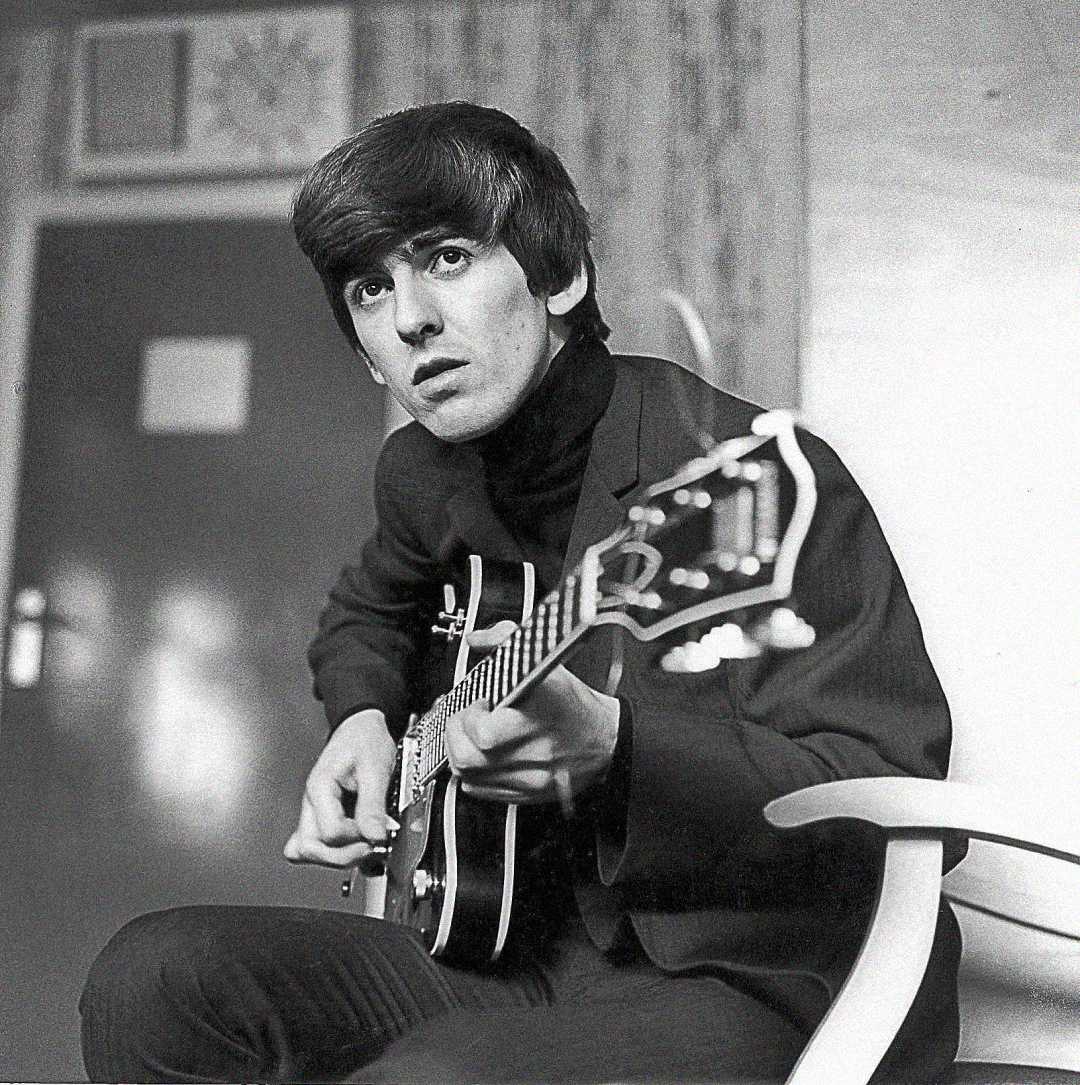 A young man with dark hair sits on a chair, playing an electric guitar and looking slightly upward. He wears a dark suit and turtleneck. The setting appears to be indoors with a door and a wall clock in the background.