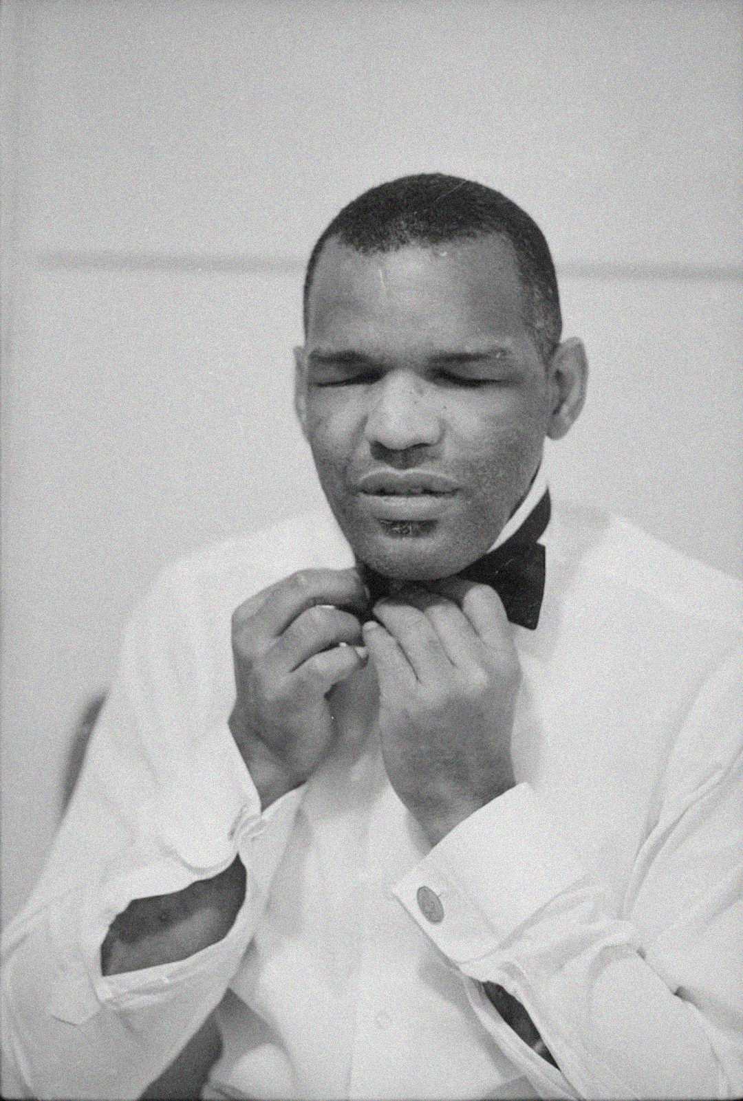 A man in a white dress shirt adjusts a black bow tie, eyes closed, in a black and white photo. The image has a vintage, classic feel.