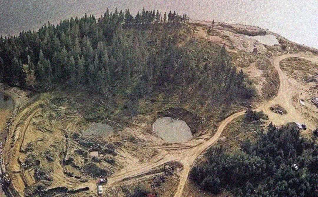 Aerial view of a forested area bordered by water, with dirt roads, clearings, and several large pits filled with water scattered throughout the landscape.