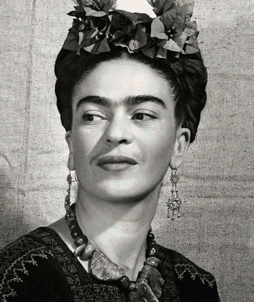 Black-and-white portrait of a woman with dark braided hair adorned with flowers, wearing bold earrings and a chunky necklace, looking slightly to the side against a plain fabric background.