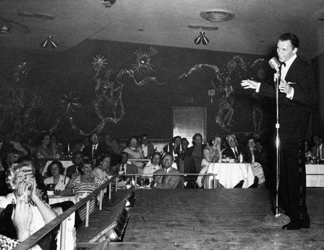 A man in a suit sings into a vintage microphone on stage at a nightclub, while a seated audience watches and smiles. The background features decorative wall art and round tables with tablecloths.