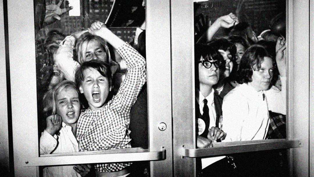 A group of excited children and teenagers press against glass doors, some with fists raised and shouting, as they try to get inside a building. The black-and-white photo captures a moment of intense anticipation and energy.