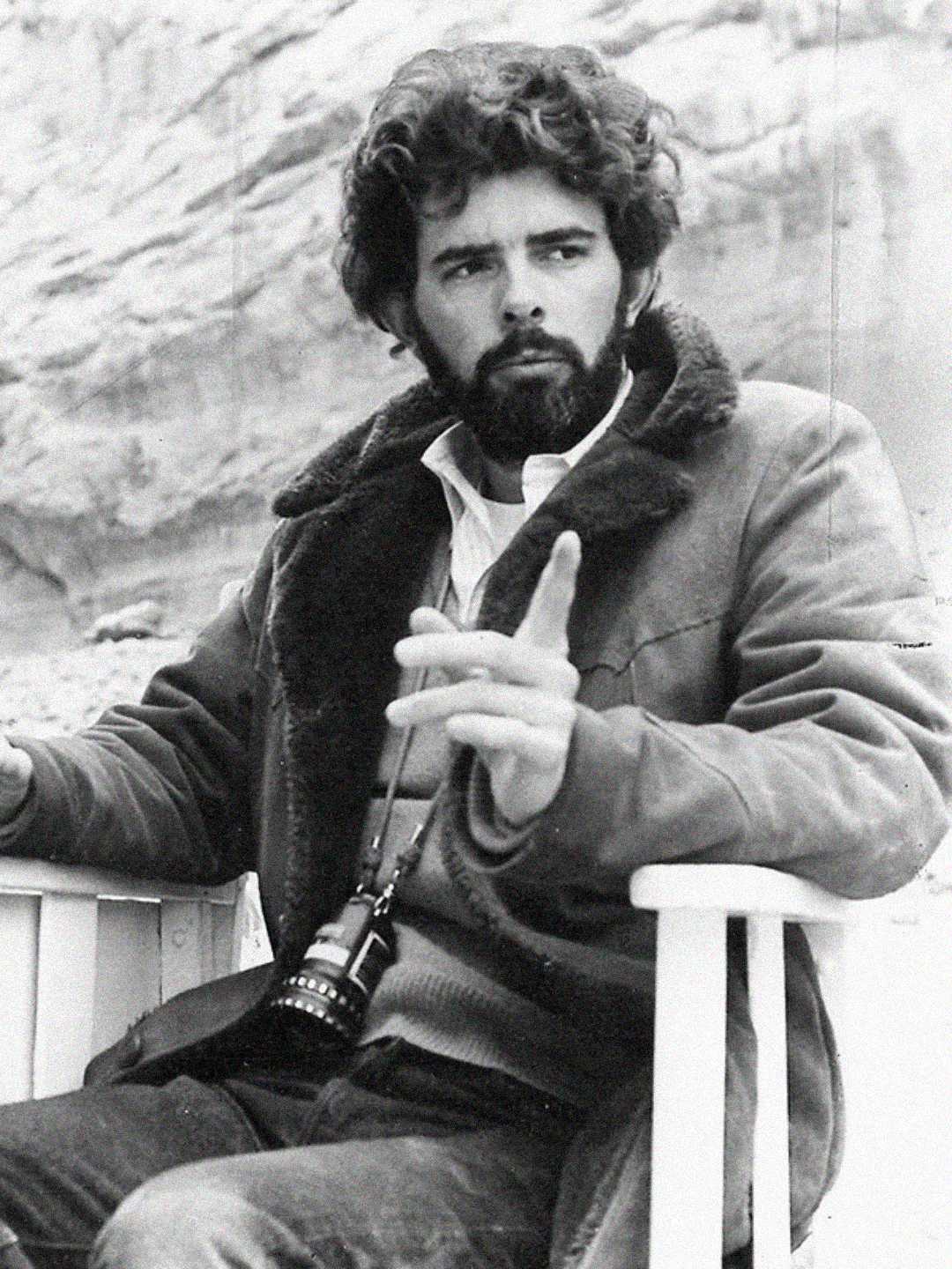 A bearded man with curly hair sits in a chair outdoors, wearing a shearling-lined jacket and holding a pair of binoculars around his neck. He gestures with one hand, with a rocky background behind him.