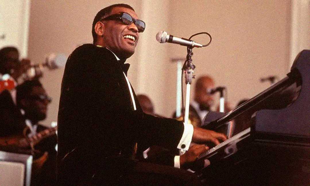 A smiling man in sunglasses and a tuxedo plays piano and sings into a microphone on stage, with other musicians blurred in the background.