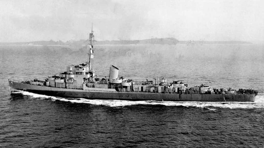 A black and white photo of a military warship sailing on the water, viewed from the side. The ship has a tall mast, smokestack, and various equipment on deck. Land is faintly visible in the distant background.