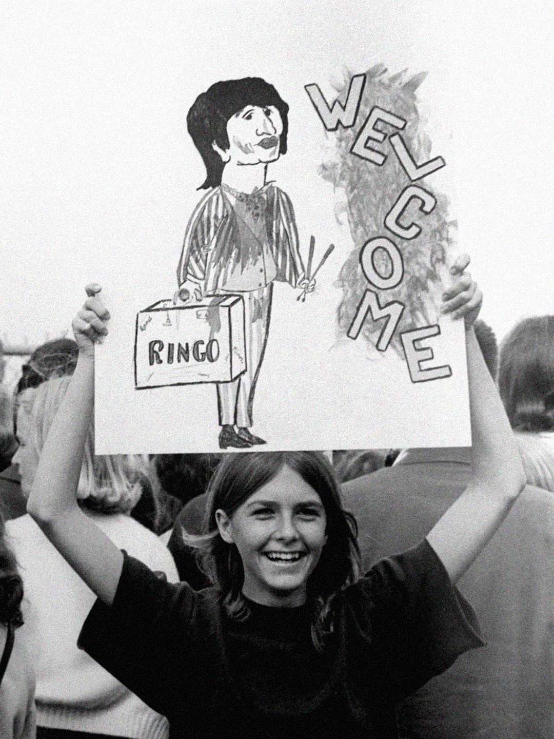 A smiling young woman in a crowd holds up a hand-drawn sign featuring a cartoon of Ringo Starr with a suitcase and drumsticks, and the word "WELCOME" in large letters.