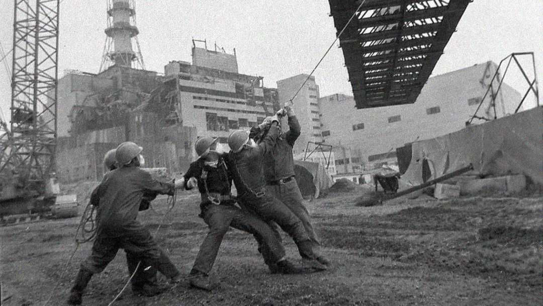Four workers in protective helmets pull a rope to guide a large metal structure near a damaged industrial building, with cranes and debris visible in the background. The scene is in black and white.
