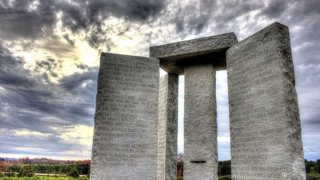 Large granite monument with tall stone slabs inscribed with text in multiple languages stands outdoors under a dramatic, cloudy sky, surrounded by a grassy landscape and distant trees.