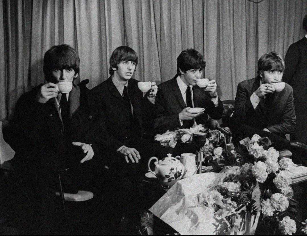 Four young men in suits sit side by side sipping tea from cups. They are surrounded by flower arrangements, a teapot, and a table covered with a tablecloth, with a curtain backdrop behind them. The image is in black and white.