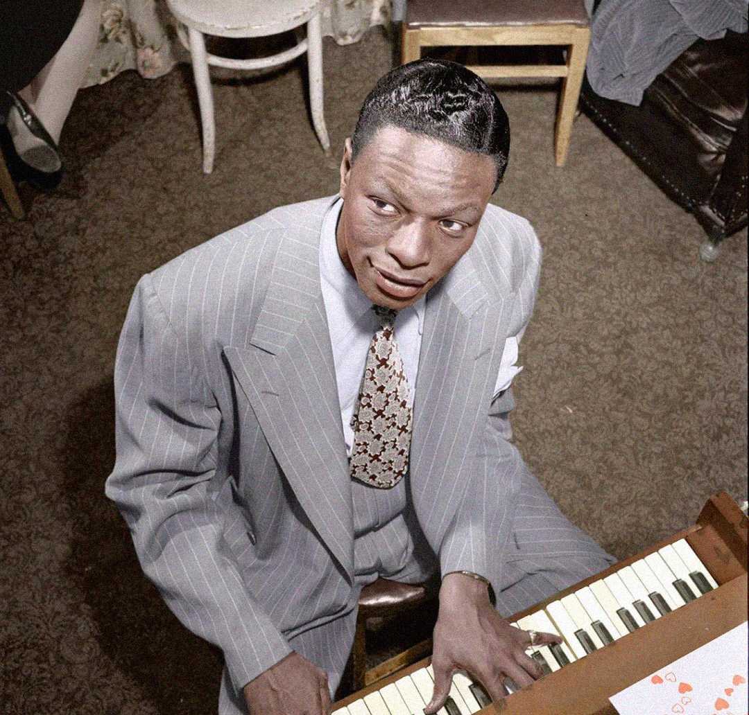 A man in a gray pinstripe suit plays the piano while looking up. He has neatly styled hair and is seated in a room with patterned carpet, a floral curtain, and wooden chairs.