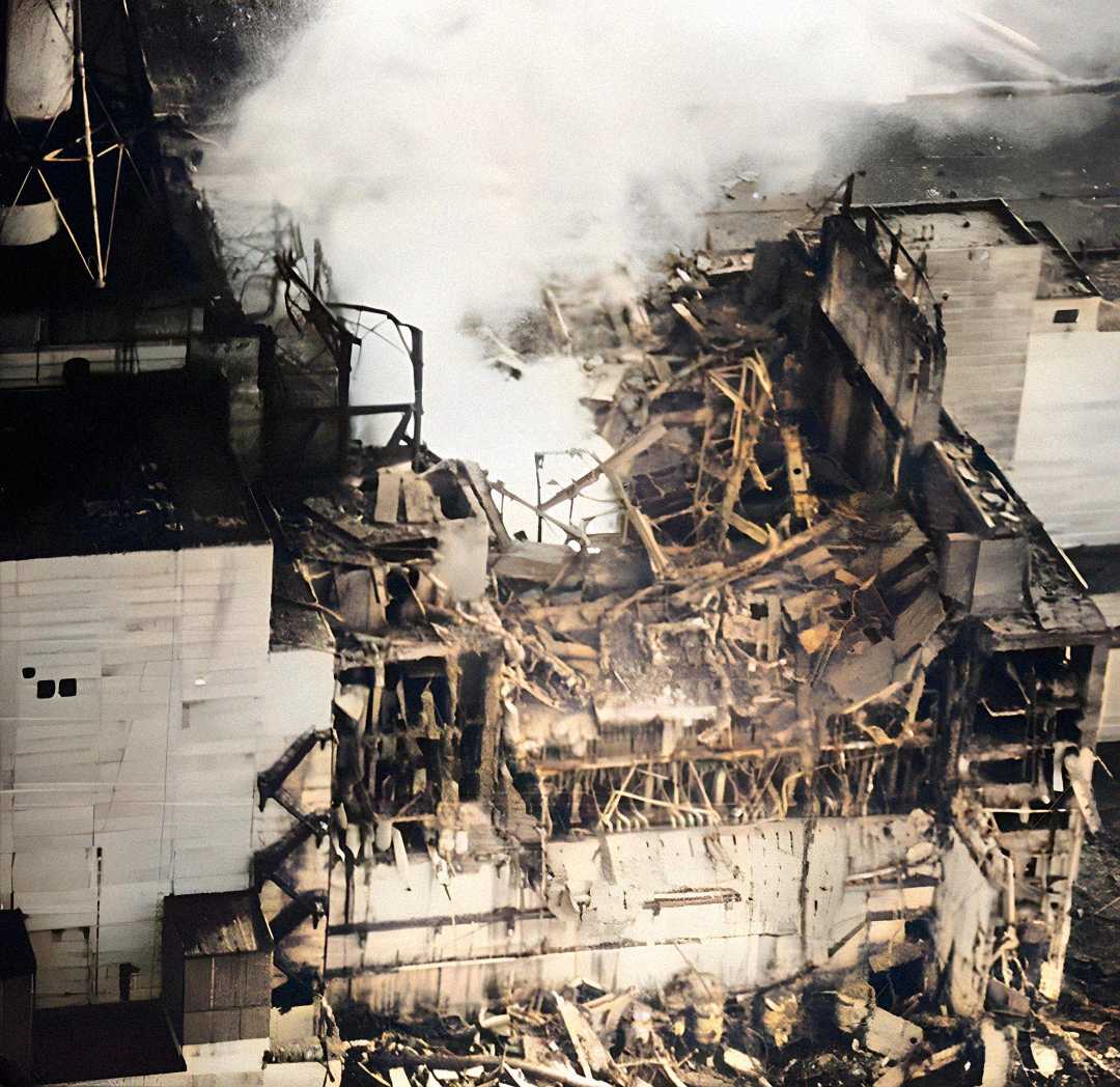 Aerial view of a severely damaged industrial building with collapsed walls, exposed steel framework, and smoke rising from the rubble, indicating the aftermath of a major explosion or disaster.