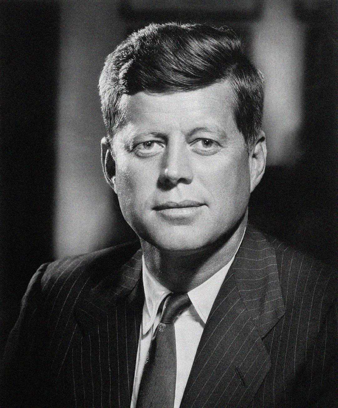 Black and white portrait of a man in a pinstripe suit and tie, with neatly combed hair, looking straight at the camera with a neutral expression against a dark background.