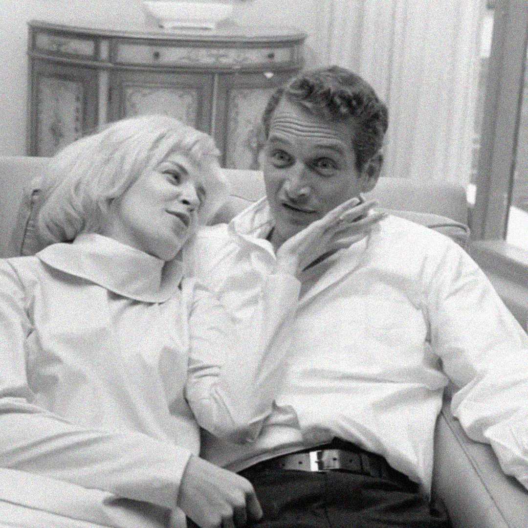 A black and white photo of a woman and a man sitting closely on a couch. The woman leans affectionately toward the man, who gestures with his hand while looking at her. Both are dressed in light-colored clothing.