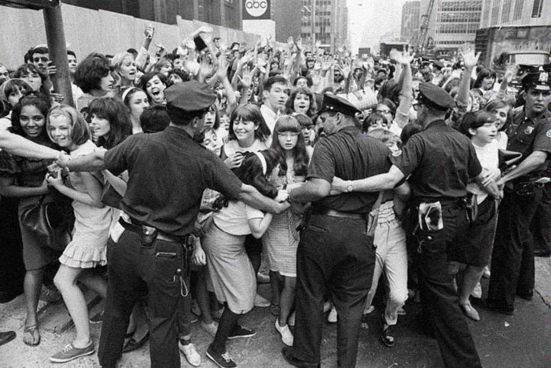 Black-and-white photo of a large crowd of excited young fans, mostly girls, being held back by uniformed police officers on a city street. The atmosphere appears energetic and frenzied.