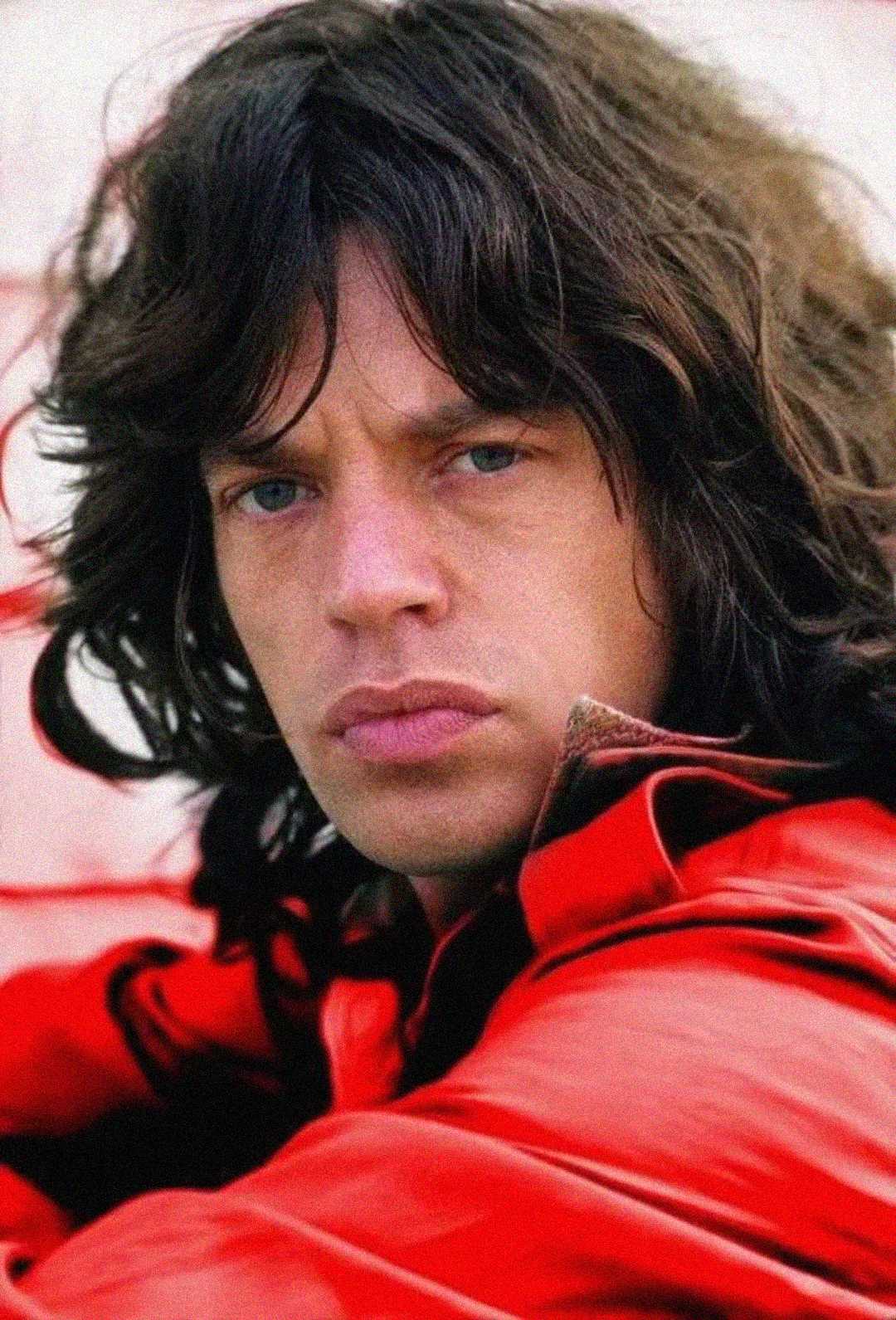 A man with long dark hair and blue eyes wears a red jacket and looks seriously at the camera against a light background.