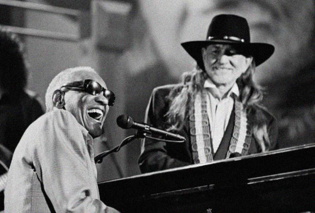 Two musicians perform together on stage; one plays the piano and sings joyfully while wearing sunglasses, and the other stands nearby in a cowboy hat, smiling. The image is in black and white.