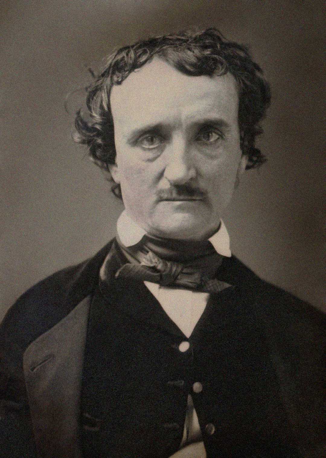 A sepia-toned portrait photograph of a man with wavy dark hair, a mustache, and intense eyes, wearing a dark jacket, waistcoat, white shirt, and a cravat. He faces the camera with a serious expression.