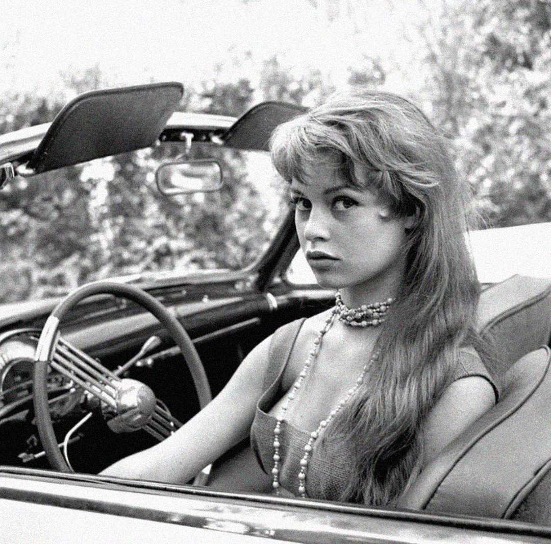 A young woman with long hair and a pearl necklace sits in the driver's seat of a classic convertible car, looking toward the camera. The photo is black and white with a blurred outdoor background.