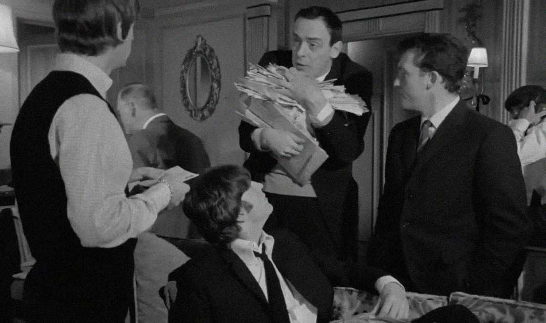 Four men in suits are gathered in a room. One man, standing and holding a large stack of papers, looks surprised or animated. Two other men stand near him, while another is seated and looking up. The setting appears formal.