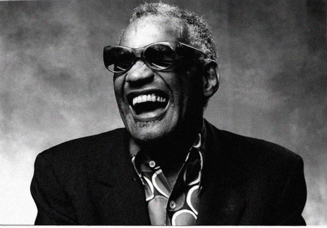 Black-and-white photo of a man wearing dark sunglasses, a suit jacket, and a patterned shirt, smiling broadly with his mouth open, showing joyful expression against a neutral background.