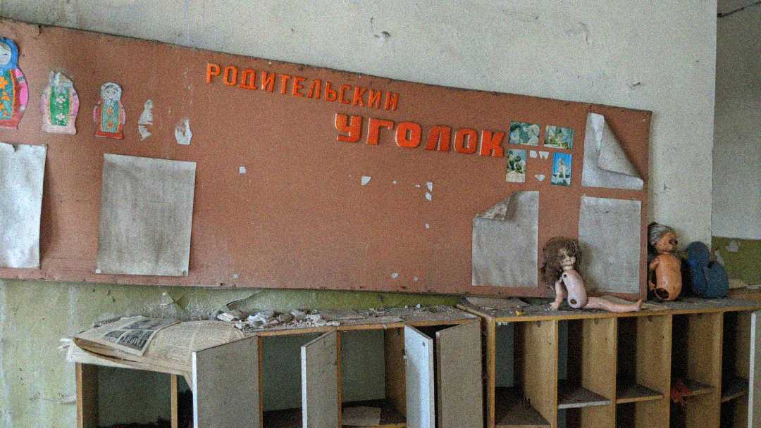 A dusty, abandoned classroom with a damaged bulletin board labeled "РОДИТЕЛЬСКИЙ УГОЛОК" in Russian, faded children's drawings, peeling paper, broken shelves, old newspapers, and worn dolls on top.