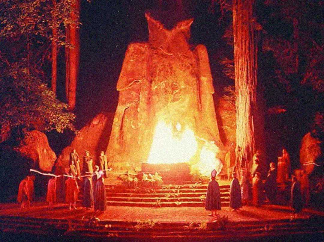 A group of people in robes stand around a large fire at the base of a giant stone owl statue, surrounded by tall trees at night, illuminated by the orange glow of the flames.