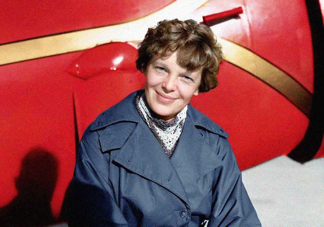 A woman with short, curly hair wearing a blue coat and patterned scarf smiles at the camera, with a red and gold airplane in the background.