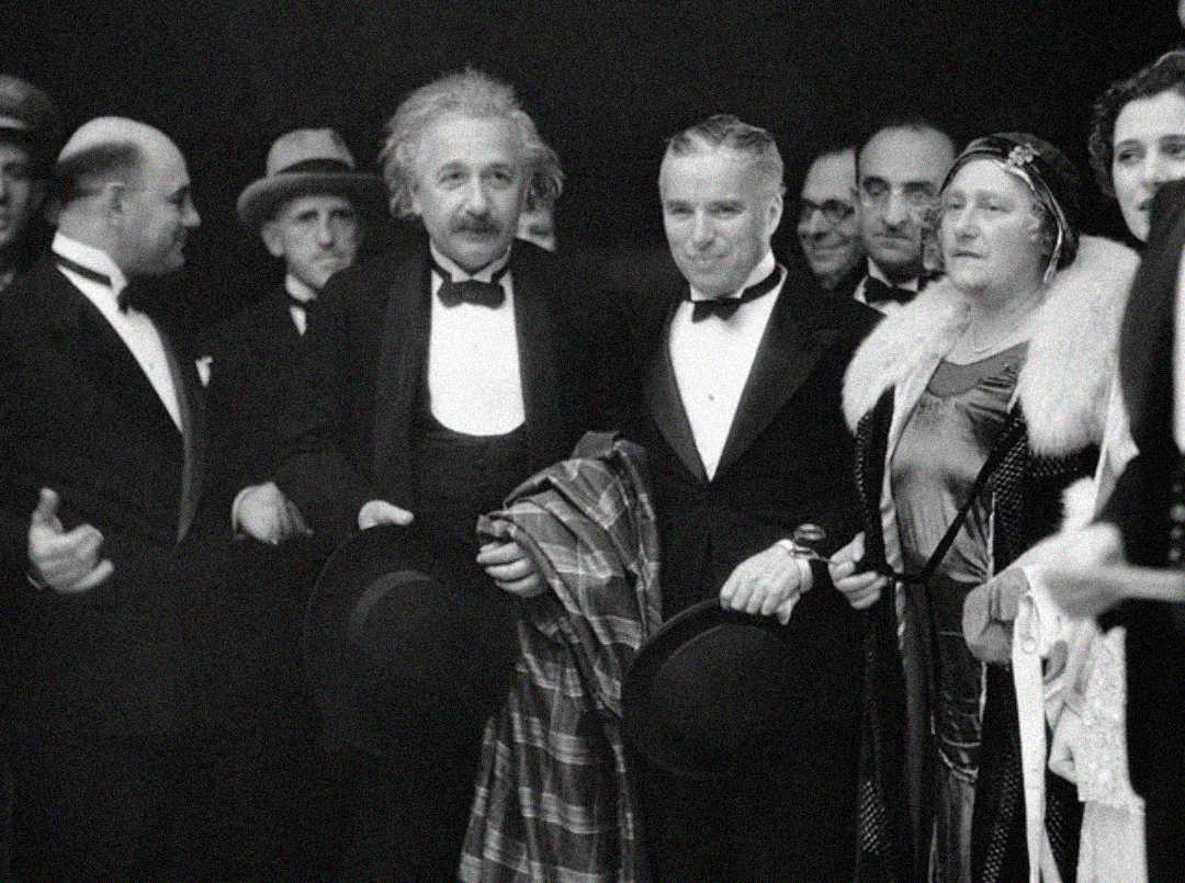 A group of people dressed in formal attire pose for a black-and-white photo; two men in tuxedos stand in the center, one holding a coat, with others gathered closely around them.