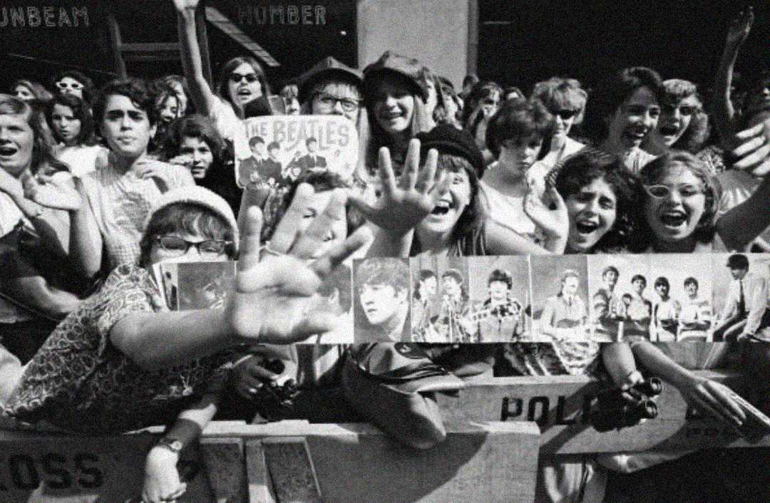 A crowd of excited fans, mostly young women, stand behind a barricade holding Beatles posters and photos, reaching out and smiling, likely at a Beatles event or concert in the 1960s.