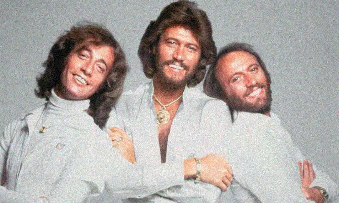 Three men with long hair and beards, dressed in white outfits, standing close together and smiling against a plain light background.