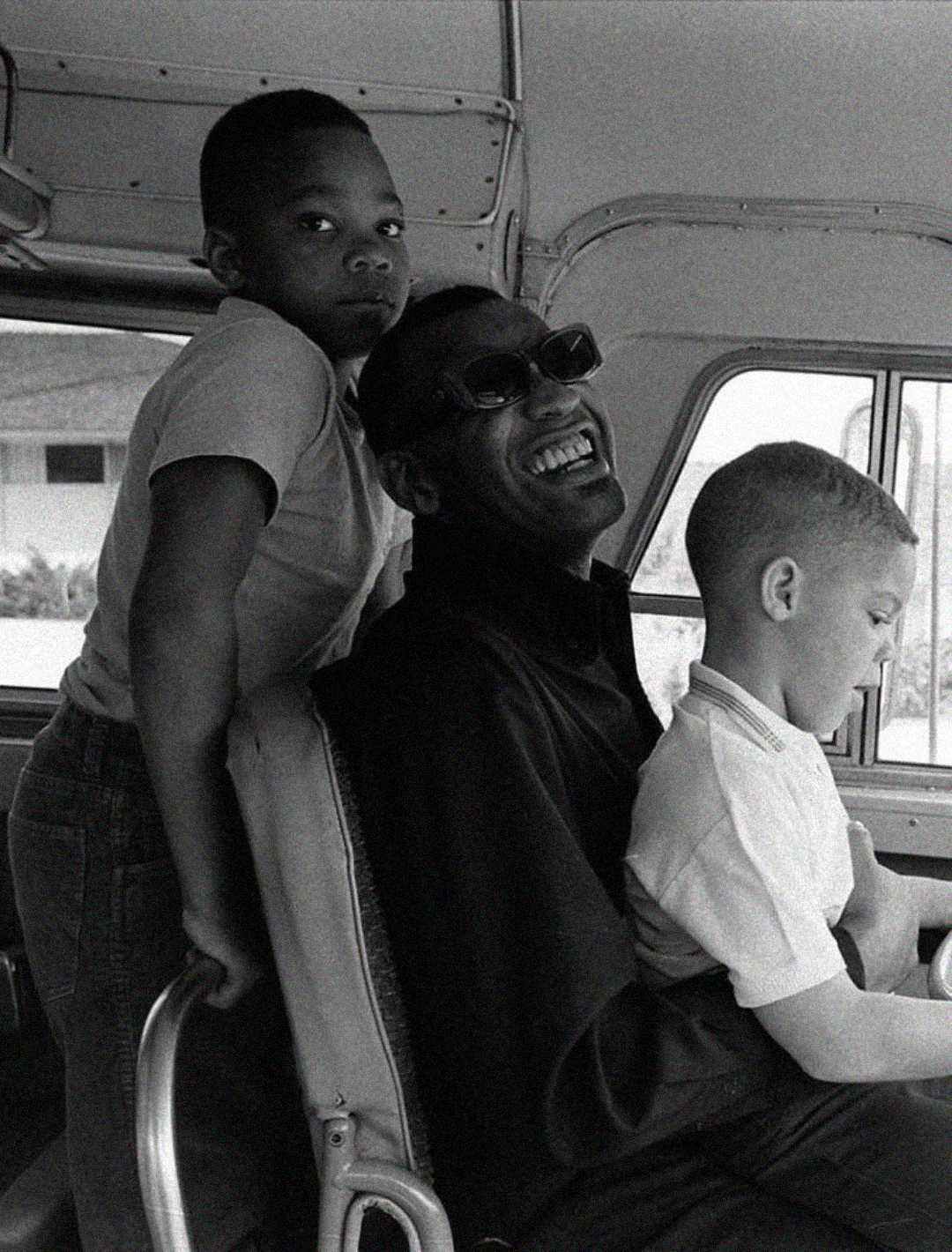 A smiling man wearing sunglasses sits on a bus seat with two young boys, one standing beside him and the other sitting on his lap. The group appears happy and relaxed.